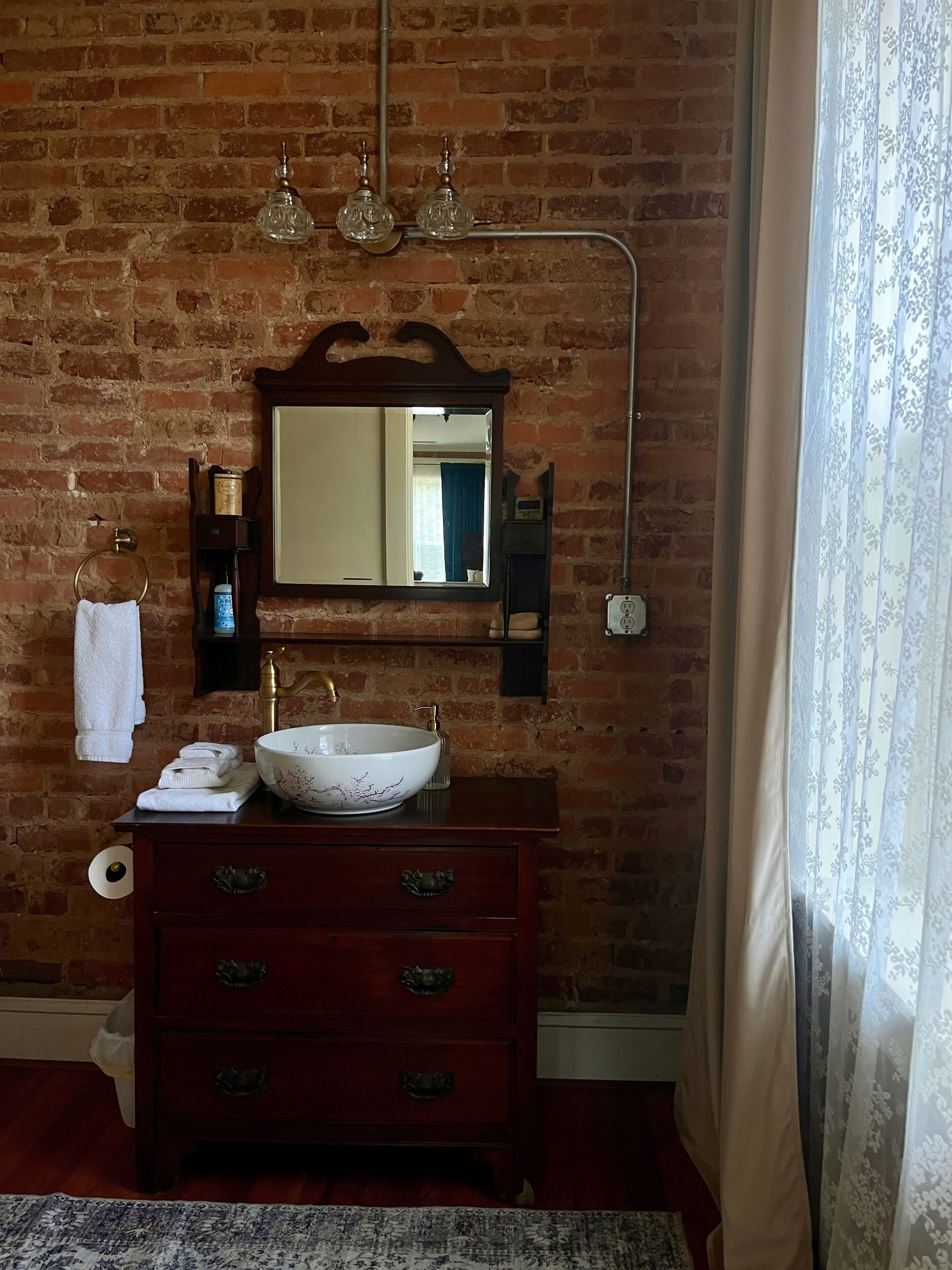 A bathroom with a brick wall, a large mirror, a white vessel sink, a wooden vanity with drawers, a towel rack, and a toilet.