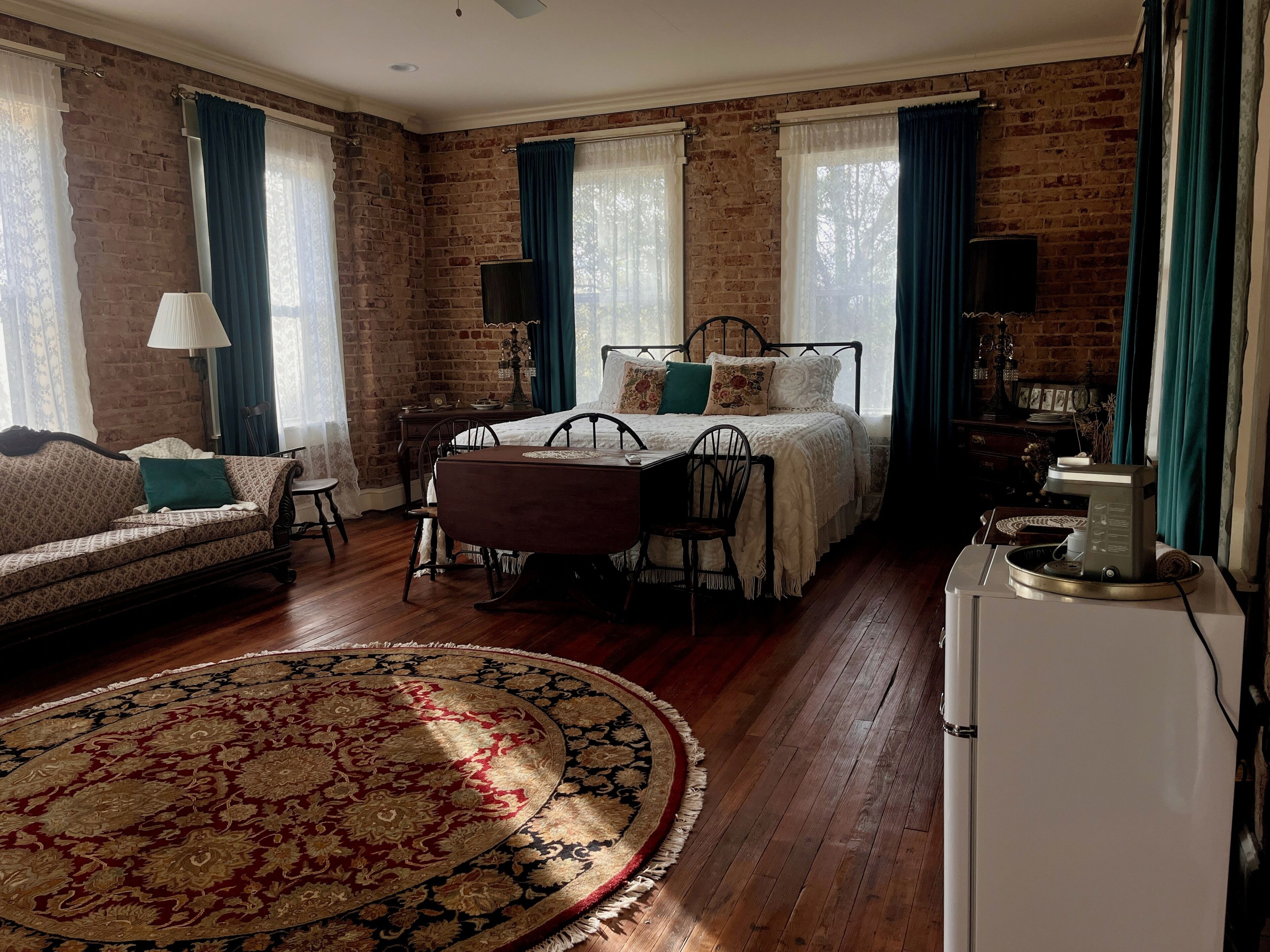 A room with a brick wall, a bed with a white bedspread, a wooden table with chairs, a coffee maker, and a large patterned rug.