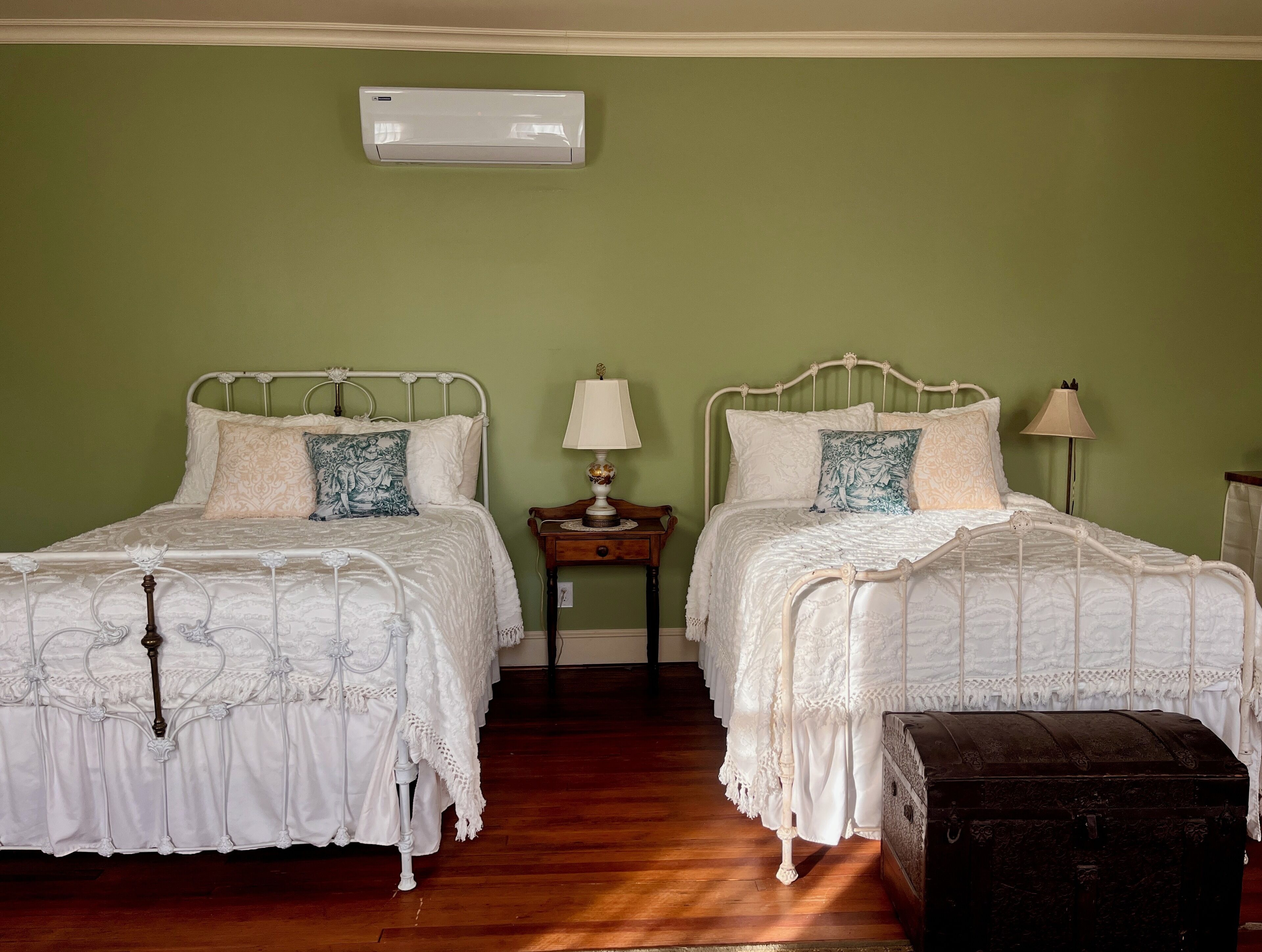 Deux lits blancs ornés de motifs complexes, une commode en bois et un climatiseur fixé au mur, le tout dans une pièce aux murs verts.