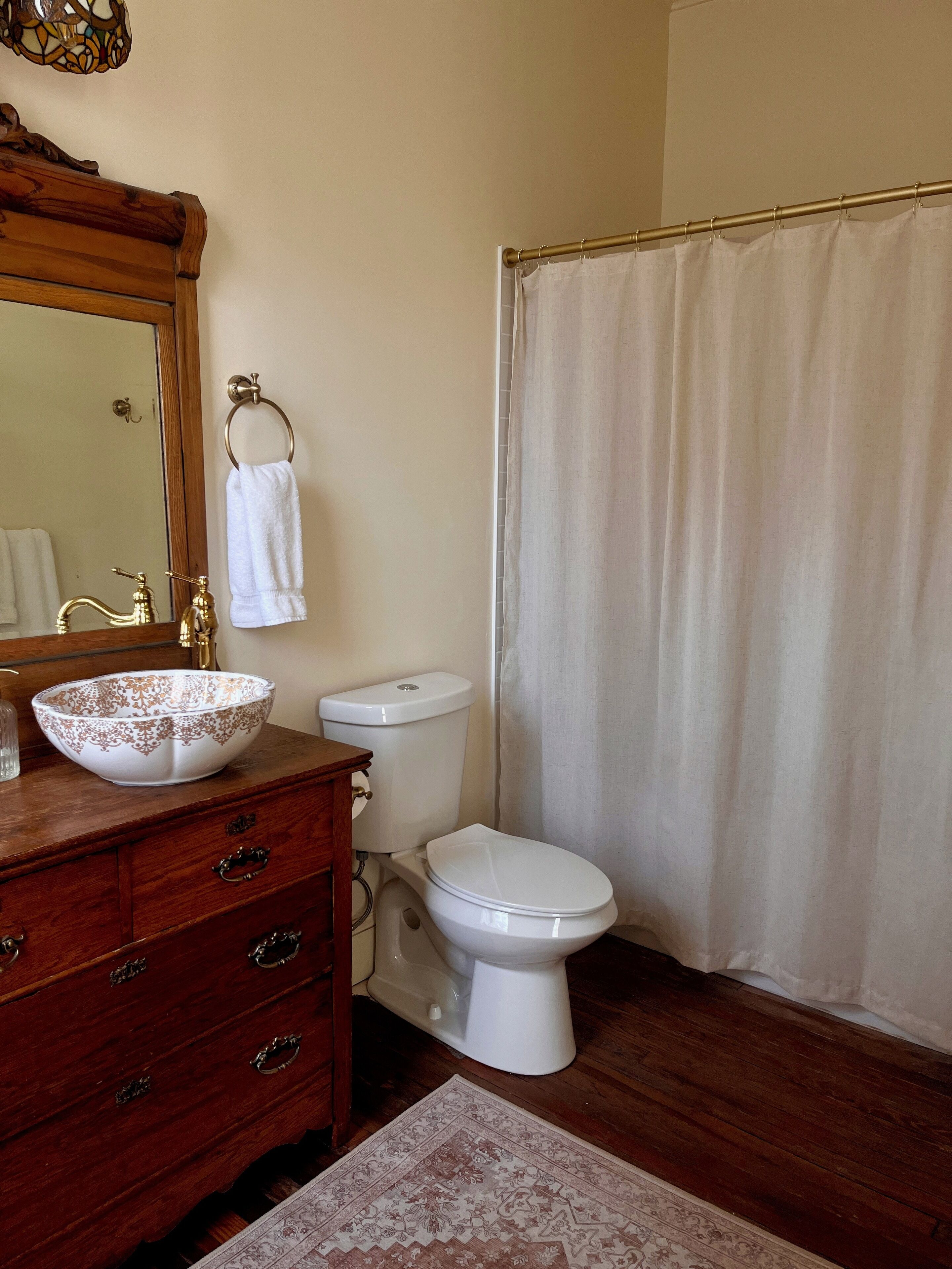 A bathroom with a wooden vanity, a white sink with a decorative bowl, a toilet, and a shower curtain.
