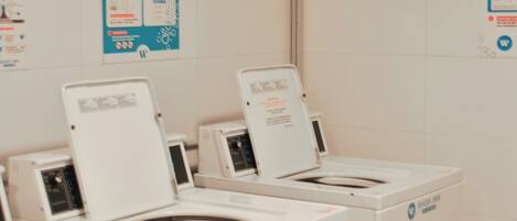 A row of white Wash Inn branded front-loading washers with control panels, set against a white tiled wall with informational posters.