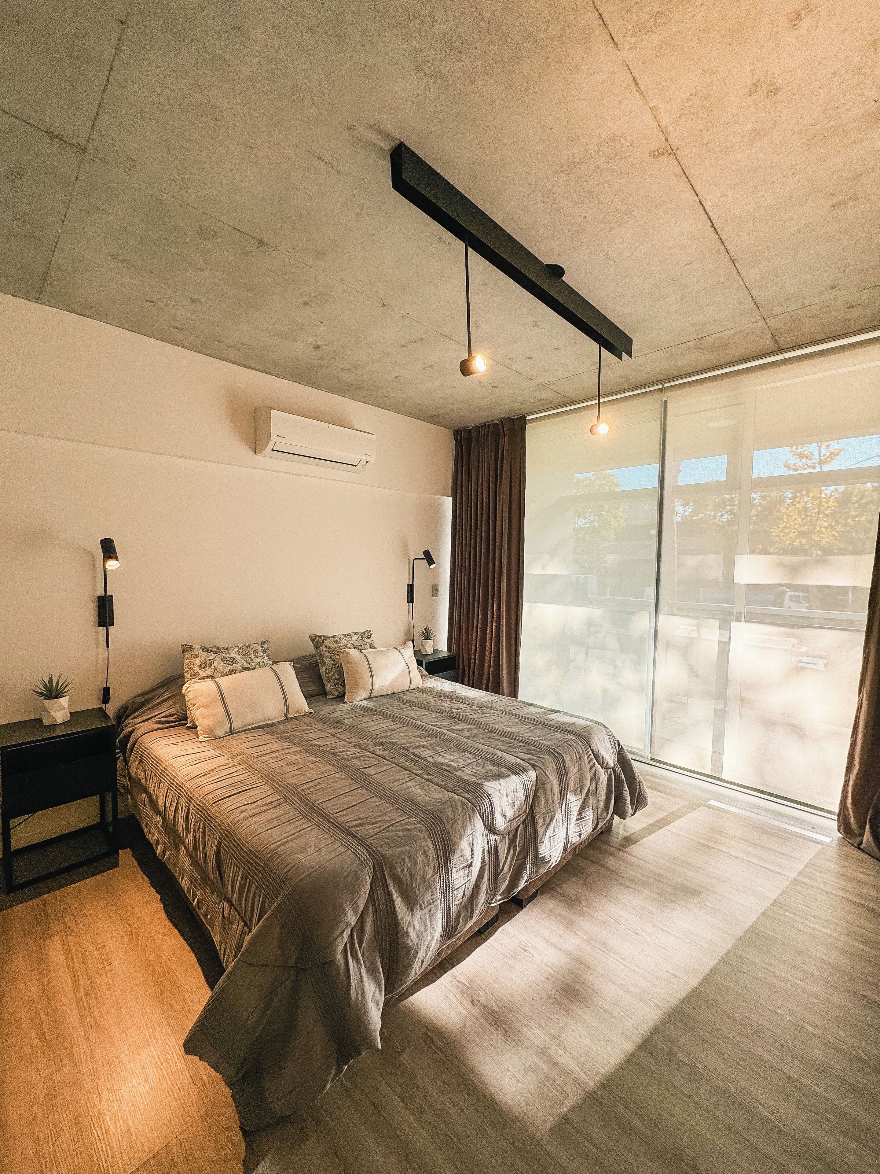 Un dormitorio moderno con una cama grande, una mesita de noche, aire acondicionado instalado en la pared y un amplio ventanal con cortinas.