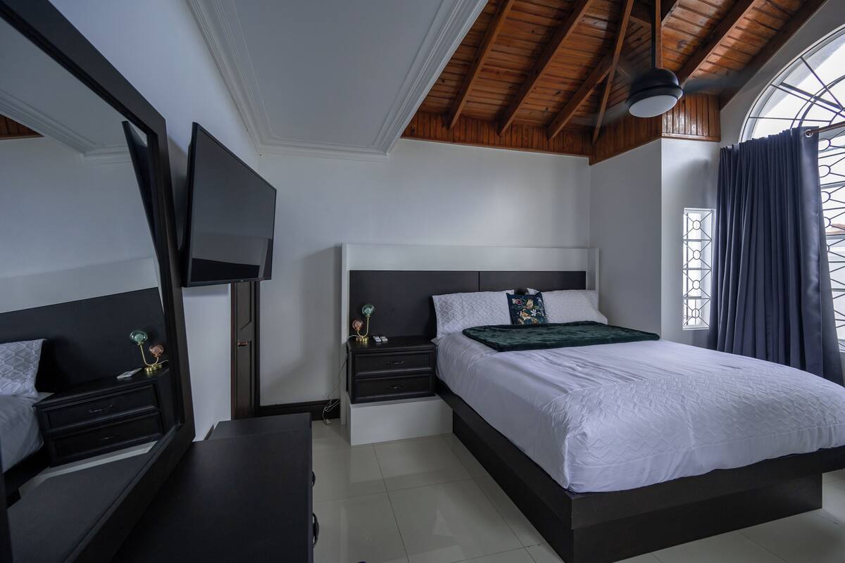 A bedroom with a large bed, a TV mounted on the wall, a nightstand, and a window with curtains.