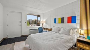 A modern hotel room with a large bed, a desk, a chair, a window with a view, and a wall with colorful panels.
