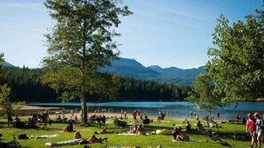 A lakeside park with people relaxing on the grass, a clear view of the lake, and mountains in the background.