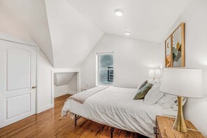 A bedroom with a sloped ceiling, a bed, a nightstand with a lamp, a window, and a door. - Spacious 4BR Duplex · 5 Blks 2 Beach · Badges Incl (Asbury Park)