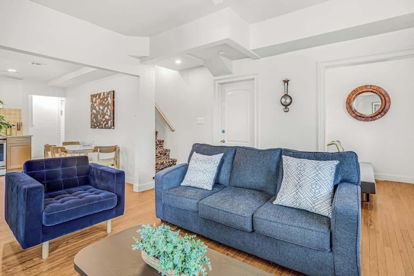 Una sala de estar con un sofá azul, un sillón azul, una mesa de centro con plantas, una zona de comedor con mesa y sillas, y una cocina con armarios y un lavabo.