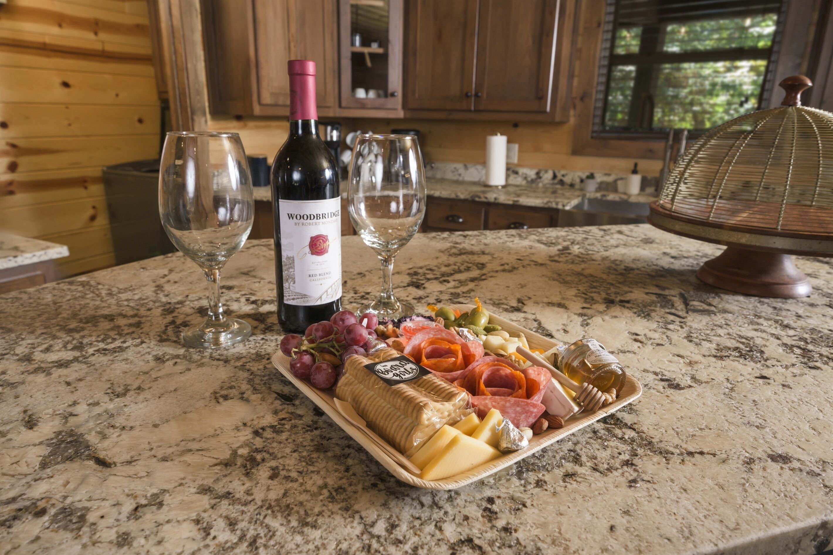 A wooden cabin interior with a marble countertop displaying a wine bottle, two glasses, and a charcuterie board with various cheeses, meats, and fruits.