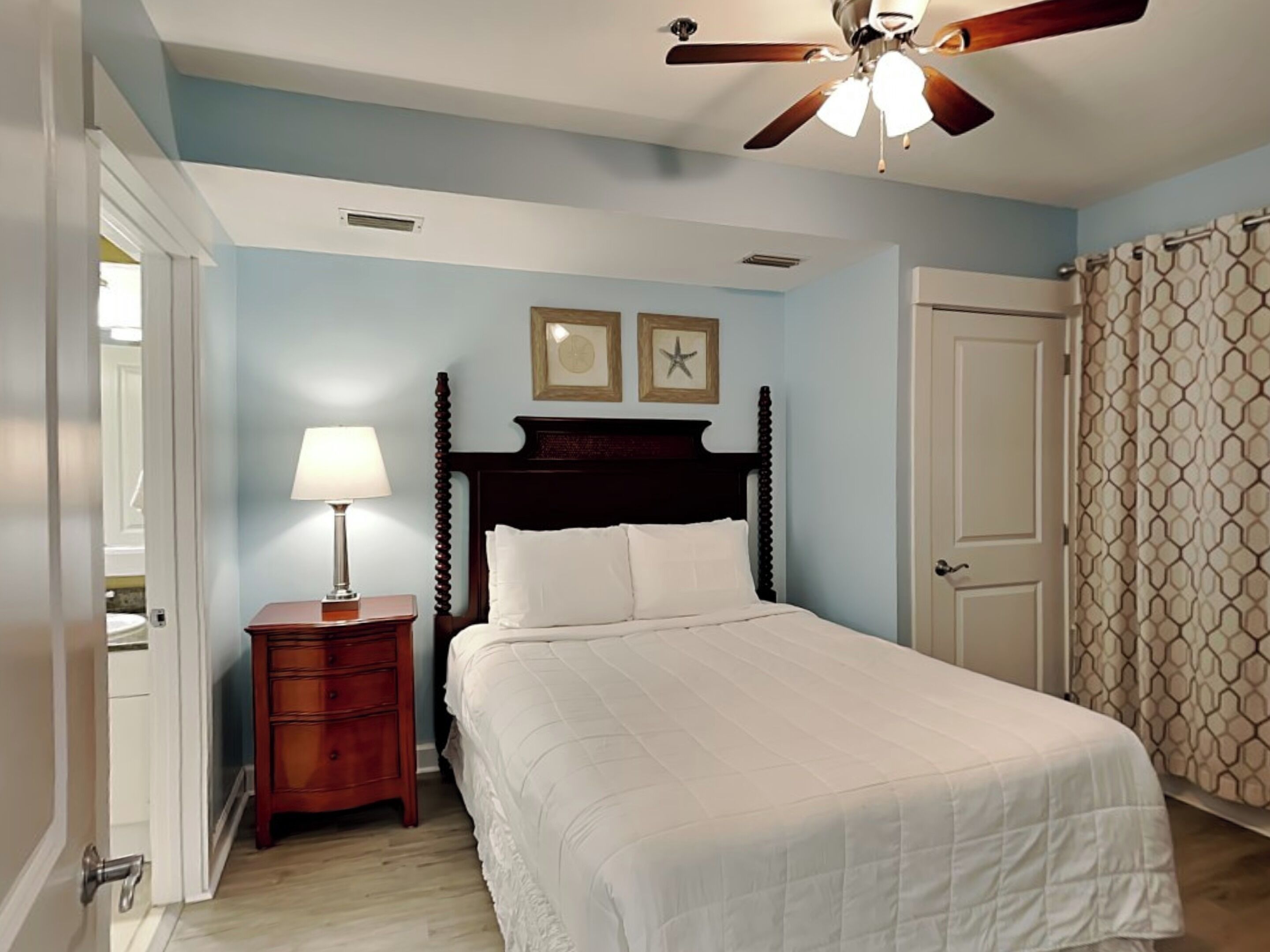 Une chambre à coucher avec un lit, une table de chevet avec une lampe, un ventilateur de plafond et une porte ornée de rideaux à motifs.