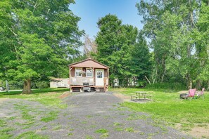 Une petite cabane en bois dotée d’un porche, entourée d’arbres et accessible par un chemin de gravier.