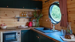 Een keuken met houten wanden, een rond raam, een spoelbak, een rode waterkoker, een fles olijfolie en een brood.