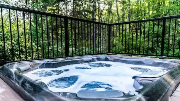 Un jacuzzi rodeado por una terraza de madera con barandilla de metal negro, con un fondo de follaje verde denso.
