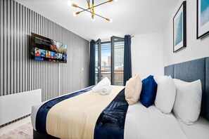 A modern hotel room with a large bed, a flat-screen TV mounted on the wall, a city view through the window, and a contemporary light fixture.