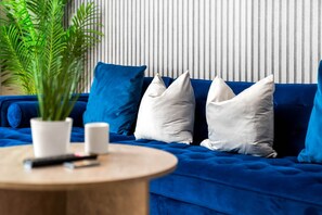 A blue sofa with white and blue cushions, a small round wooden table with a white vase and a remote control, and a green plant.