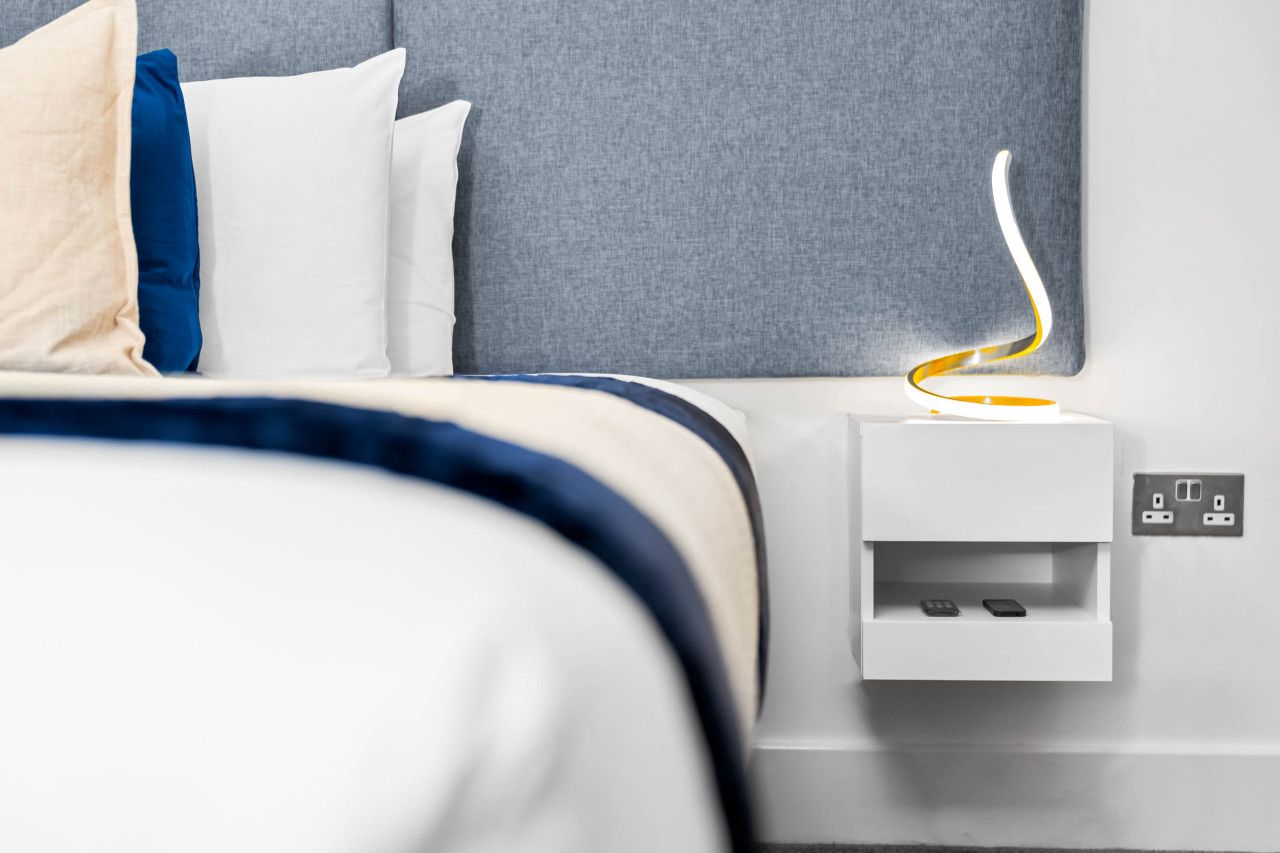 A neatly made bed with a gray headboard, white and blue bedding, and a bedside table with a power outlet and a phone charging.