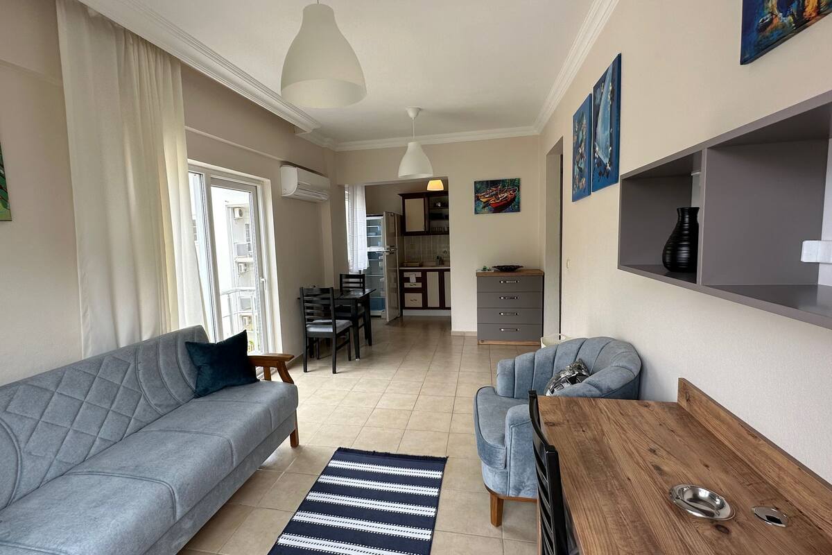 A modern living room with a grey sofa, a wooden coffee table, and a blue armchair. There is a dining area with a table and chairs, and a kitchen in the background.