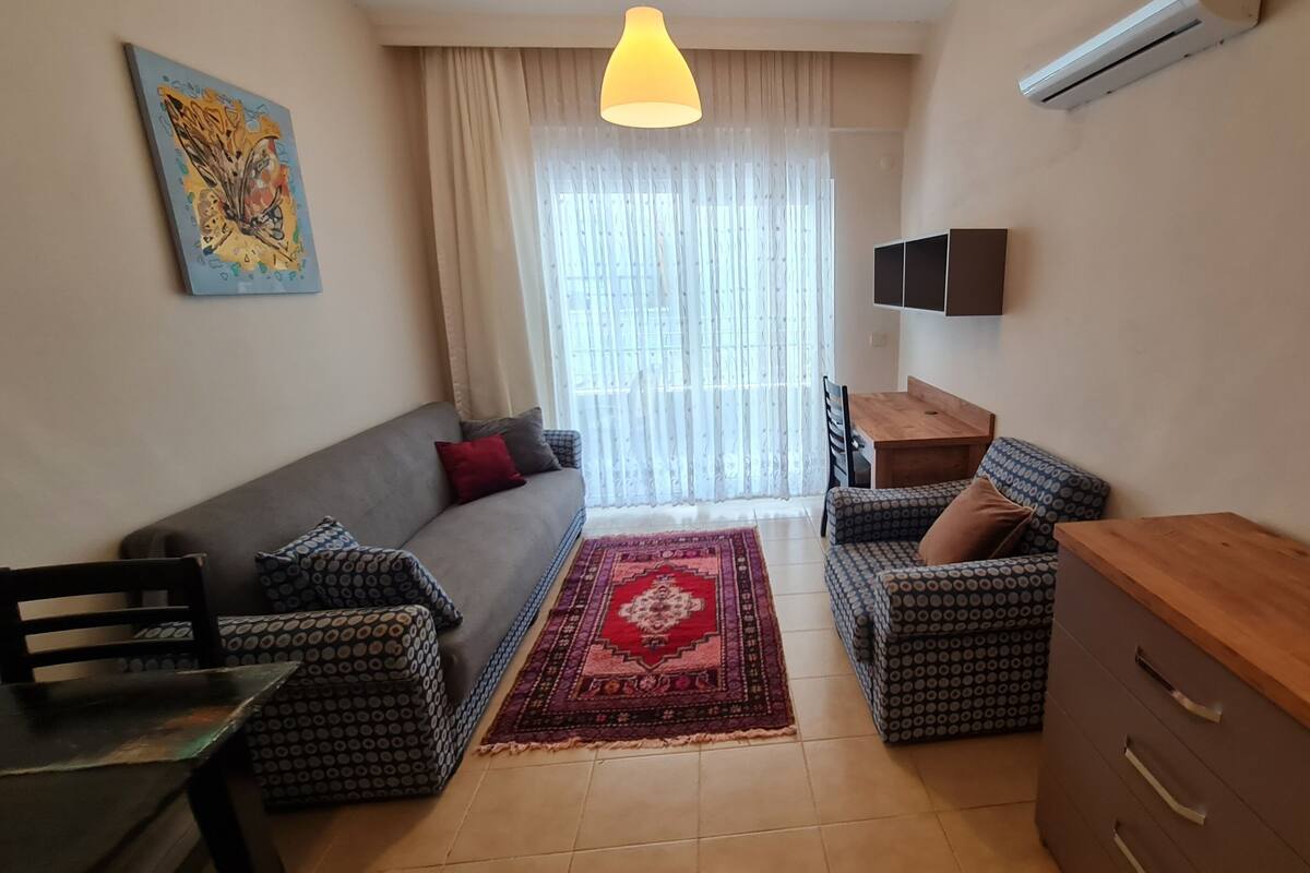 A living room with a sofa, patterned ottoman, a wooden table, a TV mounted on the wall, and a red rug.