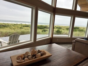 A room with large windows overlooking a grassy landscape and the ocean. There is a wooden table with a bowl of seashells. - Beauty On the Beach (Westport)
