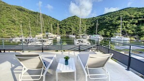 A terrace with two lounge chairs and a table, overlooking a marina with boats and lush green hills. - Marina Beach – 2 Adjacent Apartments, Marina View, Walkable Beach (Anse Marcel)