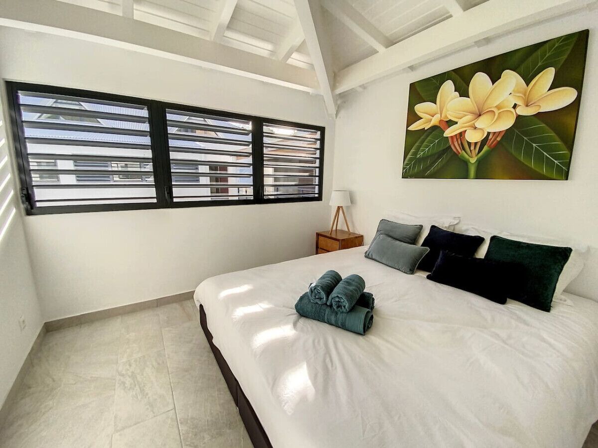 A bedroom with a large bed, a nightstand, a window with blinds, and a wall-mounted painting.