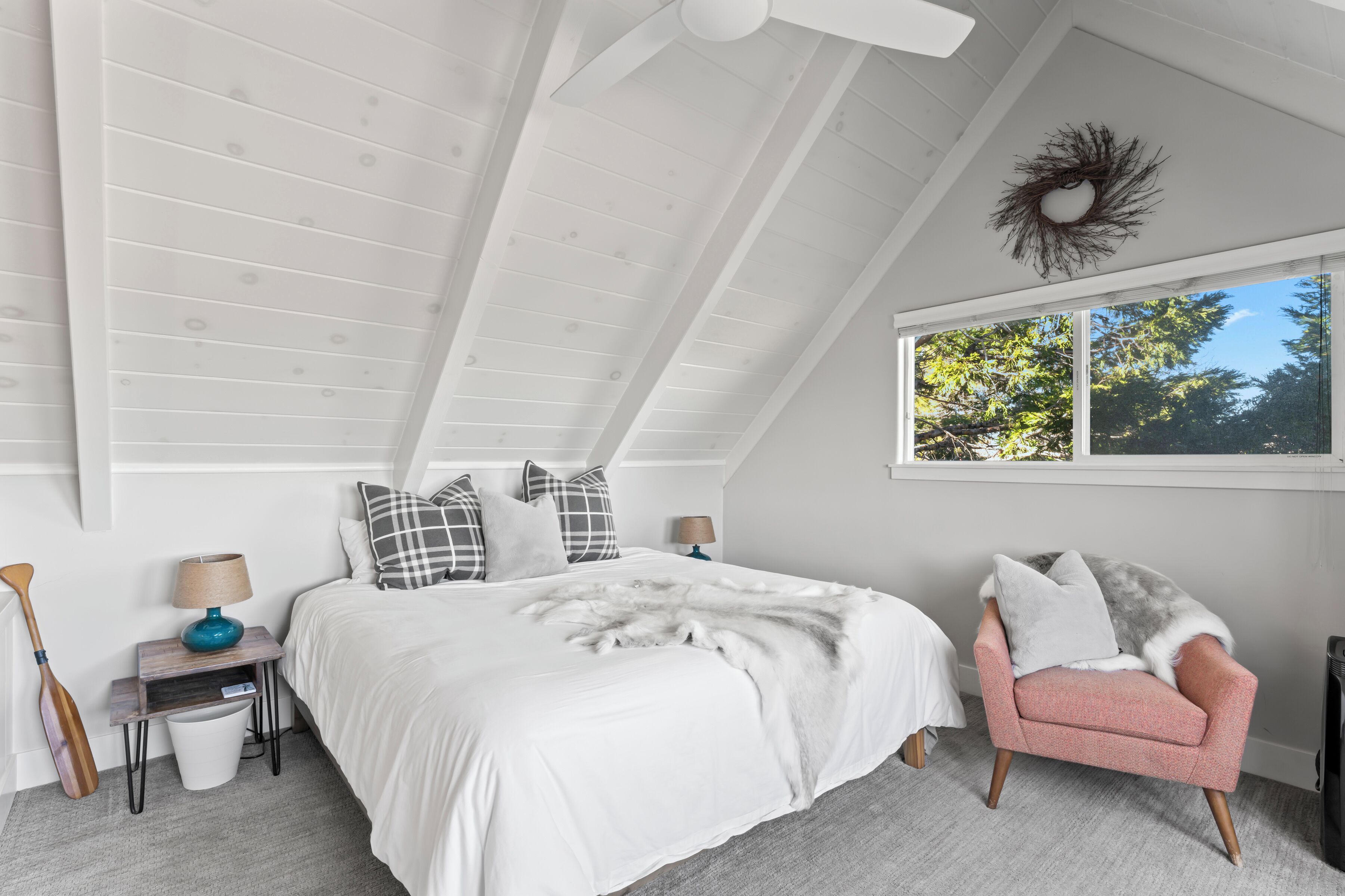 A bedroom with a sloped ceiling, a bed with a white duvet, a pink armchair, and a window with a view of trees.