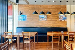 Interior de restaurante com mesas e cadeiras de madeira, um banco estofado preto e iluminação pendente.