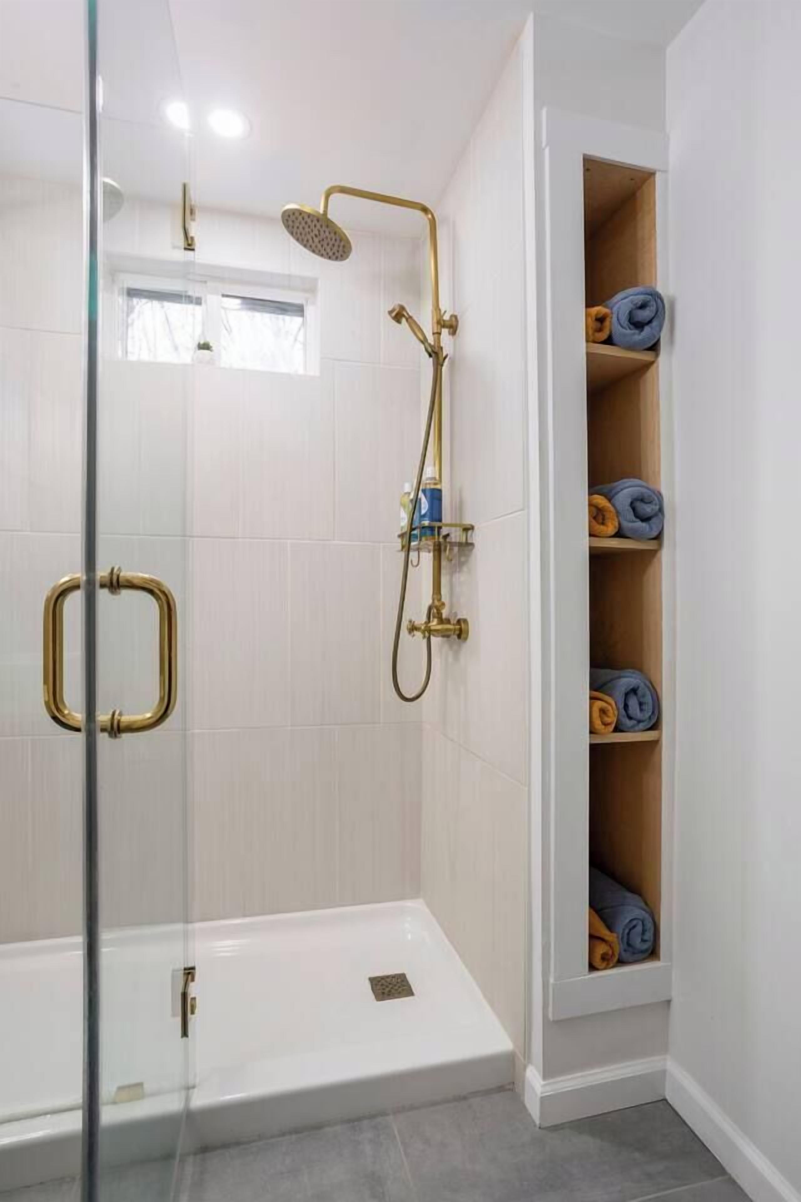 Un baño moderno con cabina de ducha de vidrio, zona de ducha con azulejos blancos y estante empotrado con toallas.