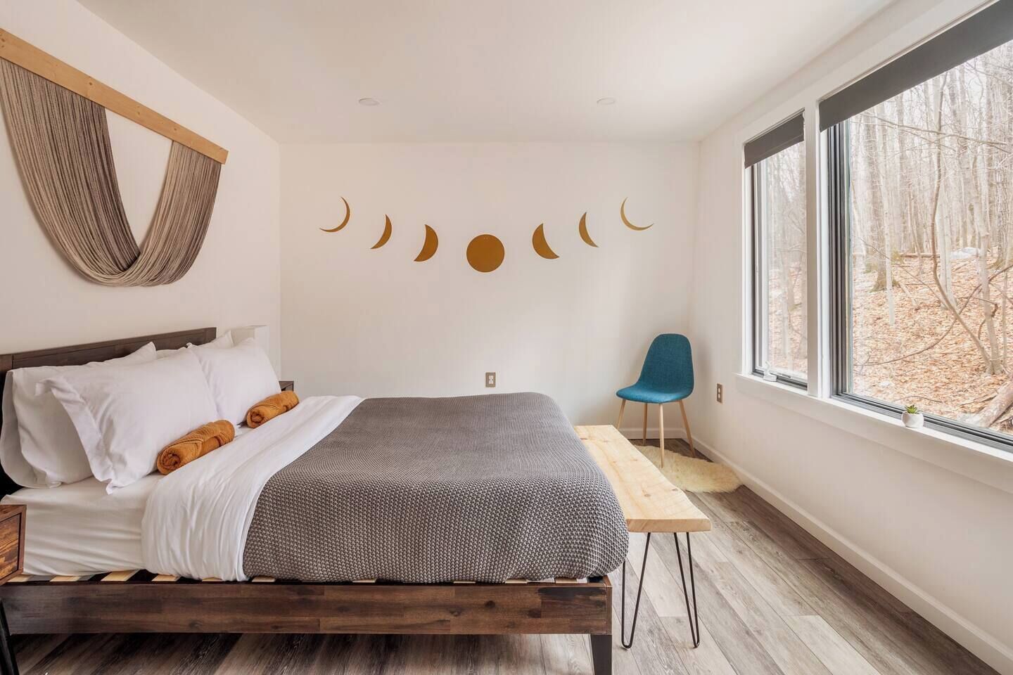 A modern bedroom with a wooden bed, a blue chair, and a large window with a view of trees.
