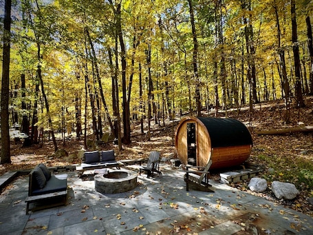 A rustic cabin with a wooden barrel entrance, stone fire pit, and outdoor seating area surrounded by a forest.