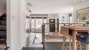 Un intérieur moderne avec un escalier menant à une salle à manger, une cuisine visible à travers une porte ouverte et une grande fenêtre qui laisse entrer la lumière naturelle.