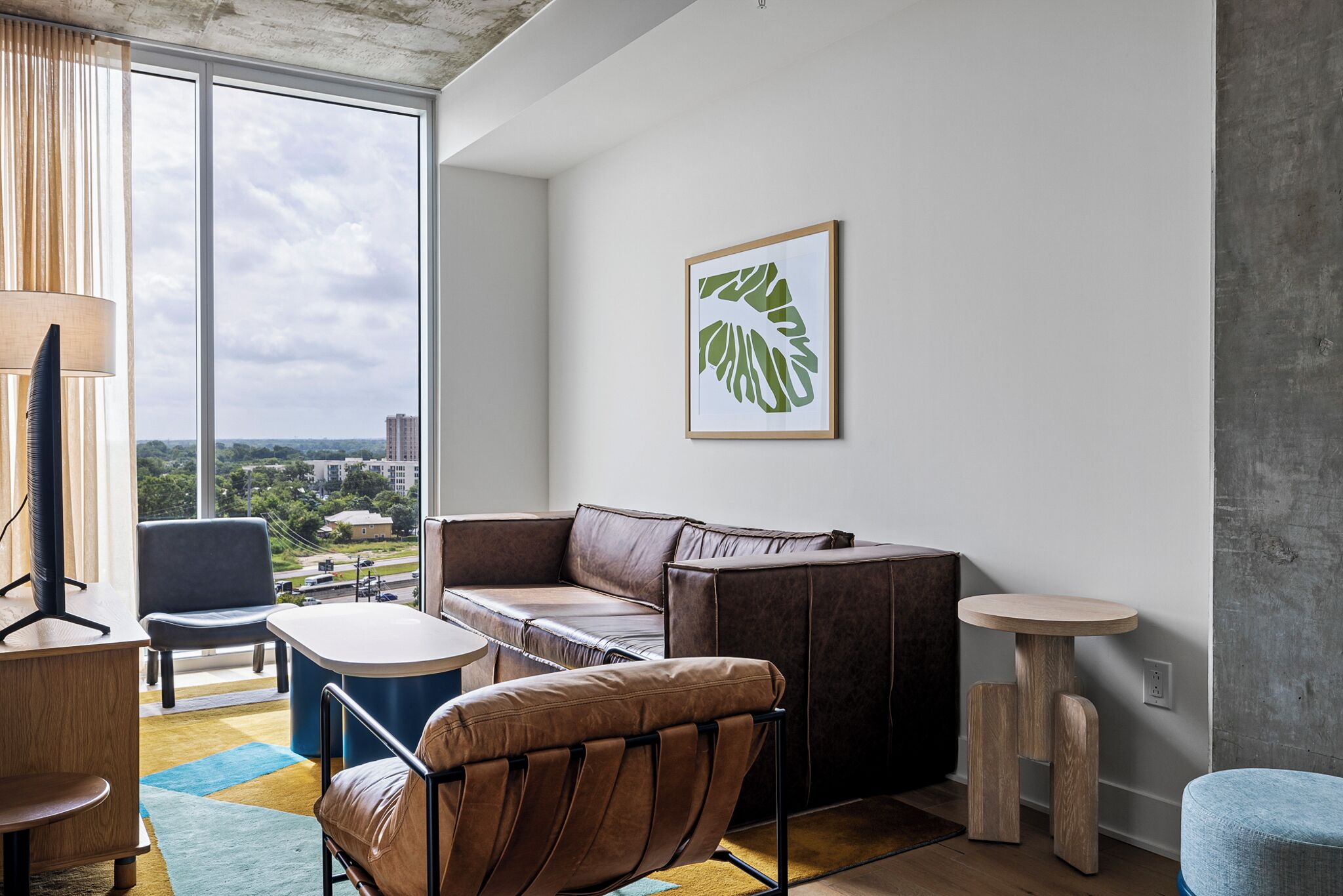 Una sala de estar moderna con un sofá de cuero, una mesita redonda y una silla. Se ve una ventana grande con vistas a la ciudad.