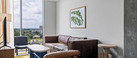 Una sala de estar moderna con un sofá de cuero, una mesita redonda y una silla. Se ve una ventana grande con vistas a la ciudad.