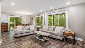A spacious living room with a large gray sectional sofa, a coffee table, and a wooden floor. There are large windows with curtains, a chandelier, and a dining area visible in the background. - Big Red! Modern clubhouse with Hot Tub! Walking distance to Castle - 4 bedrooms (Stanwood)
