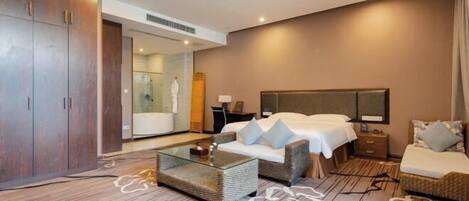 A hotel room with a large wardrobe, a glass coffee table, two sofas, a bed, and a bathroom visible through a glass door.