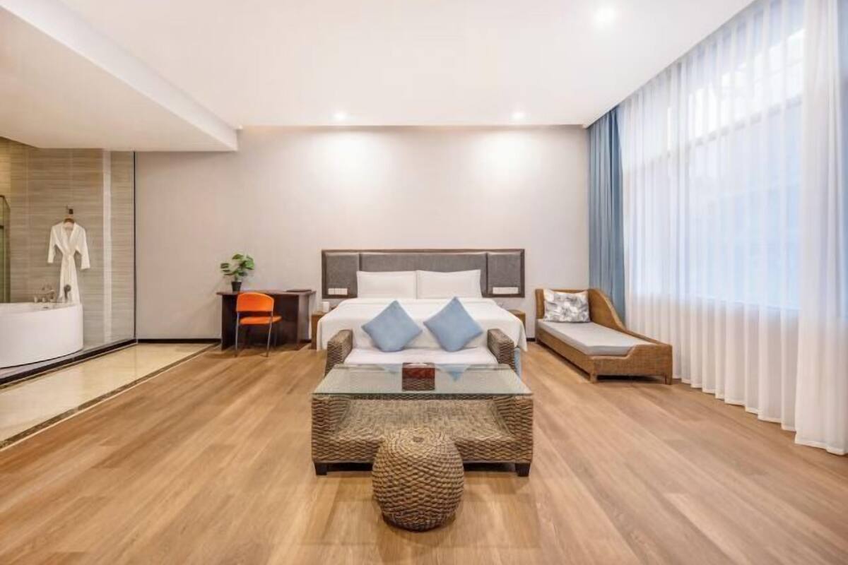 Habitación de hotel moderna con cama, una mesita de centro de cristal, un reposapiés de mimbre, una mesita auxiliar con una planta y vistas al exterior a través de cortinas traslúcidas.