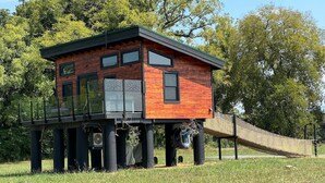 Una casa de madera sobre pilotes, con balcón, rodeada de césped y árboles.