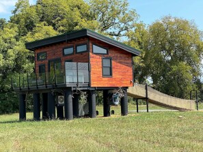 A wooden house with a balcony on stilts, surrounded by grass and trees.