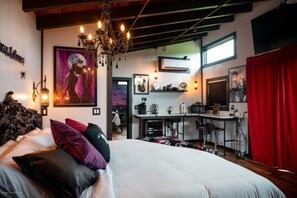 A bedroom with a large bed, a chandelier, a painting, and a kitchenette in the background.