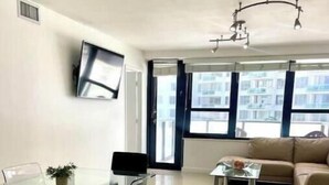 A modern living room with a glass dining table, a beige sofa, and a TV mounted on the wall. - Pretty Oceanfront Condo 2 BR w/direct access Beach (Miami)