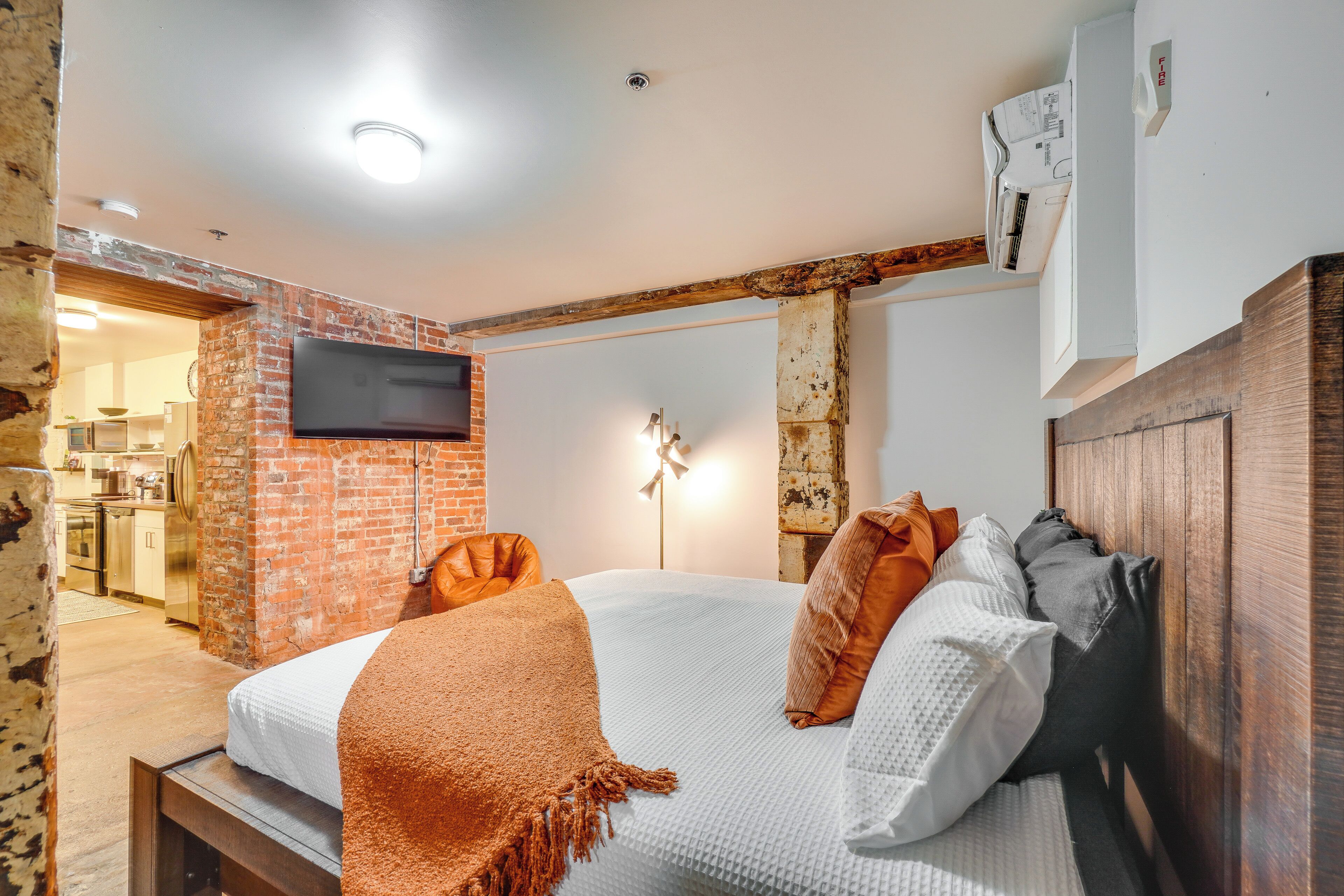 A bedroom with a brick wall, a flat-screen TV mounted above the bed, a wooden headboard, and a bedside table.