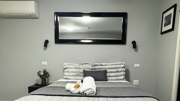 A modern bedroom with a large bed, grey bedding, a flat-screen TV above the bed, and a small bedside table with a lamp.