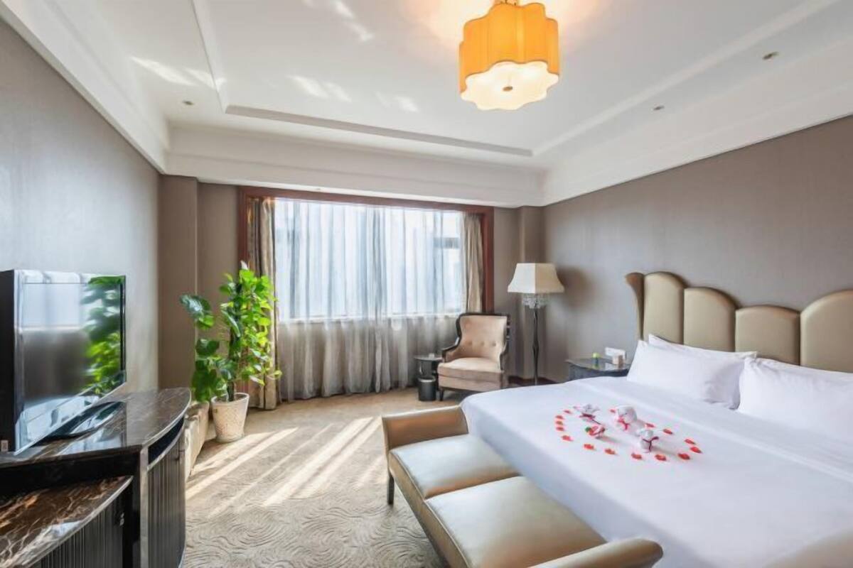 A hotel room with a bed, a TV, a desk, a chair, a lamp, and a large window with curtains.