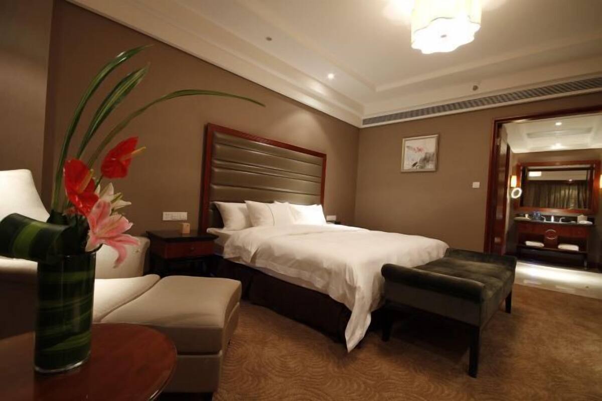 A hotel room with a bed, a nightstand, a chair, an ottoman, and a vase with flowers.