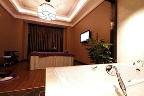 A hotel room with a large bathtub, a massage table, a television, and a potted plant.