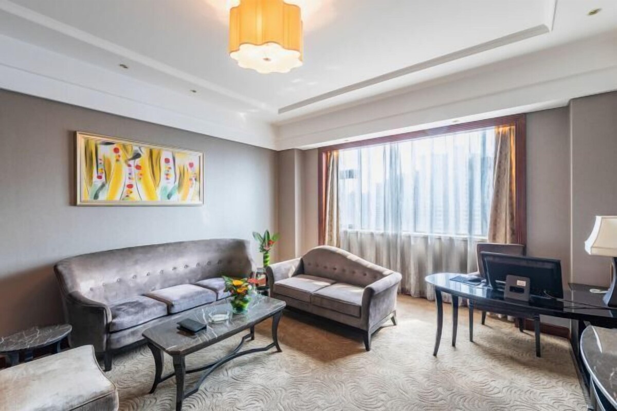 A modern hotel room with a sofa, armchair, coffee table, and a desk with a computer.