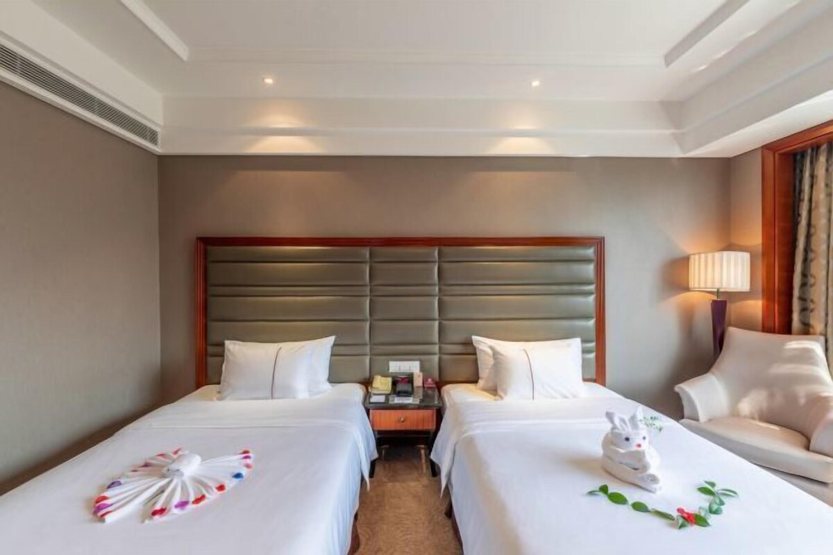 A hotel room with two beds, a wooden headboard, a bedside table with a lamp, and a chair.