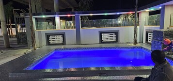 Una piscina exterior con un borde azul iluminado, rodeada por un solárium de concreto y una barandilla de vidrio.