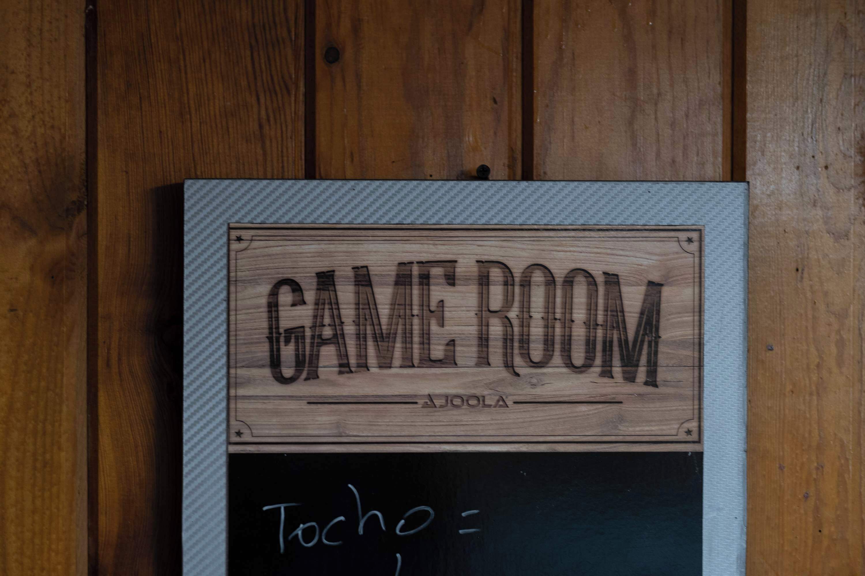 Een houten bord met daarop 'GAME ROOM' en 'AJOLA', bevestigd aan een muur.