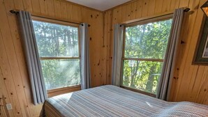 A wooden cabin room with a bed, two windows, and a view of trees.