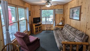 A wooden cabin interior with a sofa, a television, a dining table, and a ceiling fan.